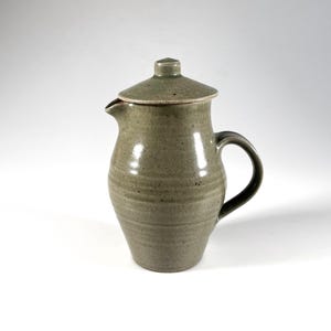 May include: A handmade stoneware jug with a lid. The jug is a muted green-grey colour with a dark handle and spout. The lid has a small knob. The jug has a rounded body with horizontal ridges.