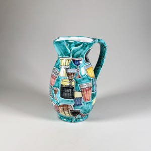 Può includere: Un'anfora in ceramica turchese con manico bianco. L'anfora è decorata con un motivo colorato di bicchieri e bottiglie di vino.