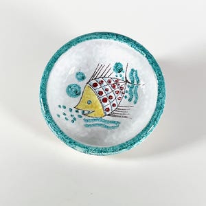 May include: A small white ceramic bowl with a turquoise rim. The bowl features a hand-painted design of a yellow fish with red and black details, surrounded by turquoise and white dots.