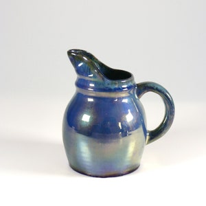 Puede incluir: Un pequeño creamer de cerámica azul con un glaseado iridiscente y un cuerpo redondeado. El creamer tiene un asa y un pico.