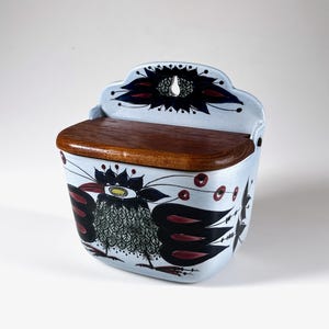 Puede incluir: Un salero de cerámica azul claro para colgar en la pared con tapa de madera. La caja está decorada con un diseño de pájaro negro y rojo. La parte superior de la caja tiene un diseño decorativo con un agujero para colgar.