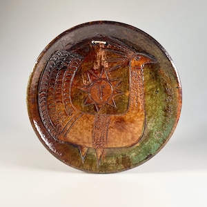 May include: A round ceramic wall plate featuring a glazed finish in shades of brown, green, and amber. The surface displays an embossed design of a stylised bird with a long neck and a sun motif with a face, along with incised decorative patterns.