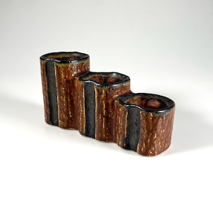 May include: A three-section ceramic candle holder. The holder is brown with black vertical stripes and a glossy finish. Each section's interior is dark brown. The design is reminiscent of a tree trunk.