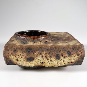 Puede incluir: Un recipiente de cerámica cuadrado hecho a mano con una abertura circular marrón oscuro. El exterior presenta una superficie texturizada y porosa con tonos beige, marrón y negro. La pieza de cerámica tiene una estética orgánica única.