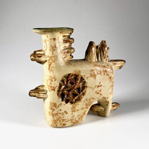 May include: A beige ceramic vase with an abstract design. The vase has a textured surface with brown speckles and a circular, patterned indentation. It has a rectangular body with protruding elements and a green-tinged rim.