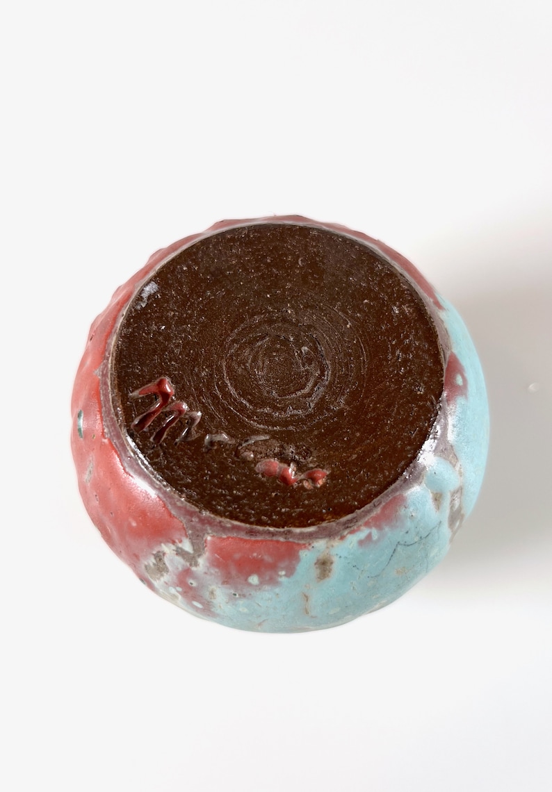May include: A small, round ceramic bowl with a brown glaze on the inside and a blue and red glaze on the outside. The bowl has a signature on the bottom.