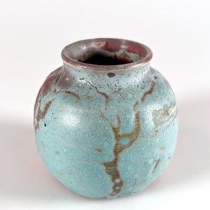 May include: A small, round ceramic vase with a light blue glaze and brown and red accents. The vase has a slightly uneven surface and a wide opening.