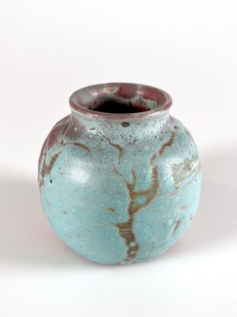 May include: A small, round ceramic vase with a light blue glaze and brown and red accents. The vase has a slightly uneven surface and a wide opening.