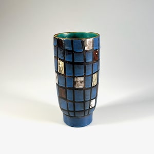 May include: A tall, blue ceramic vase with a geometric pattern of squares in shades of blue, brown, and silver. The vase has a teal interior and a gold rim. The base is a darker shade of blue.