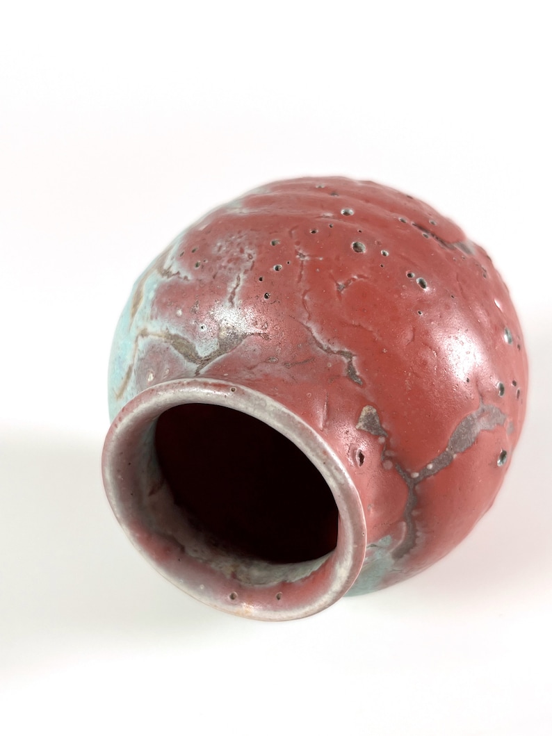 May include: A red and blue ceramic vase with a textured surface and small holes. The vase is tilted on its side, revealing the inside.