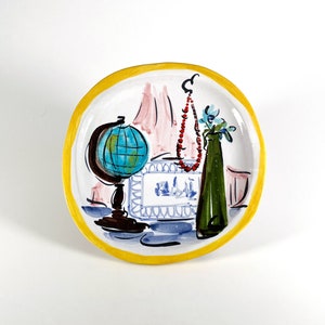May include: A ceramic plate with a yellow rim features a hand-painted design of a globe, a vase with blue flowers, and a red beaded necklace. The plate is a decorative piece with a whimsical style.
