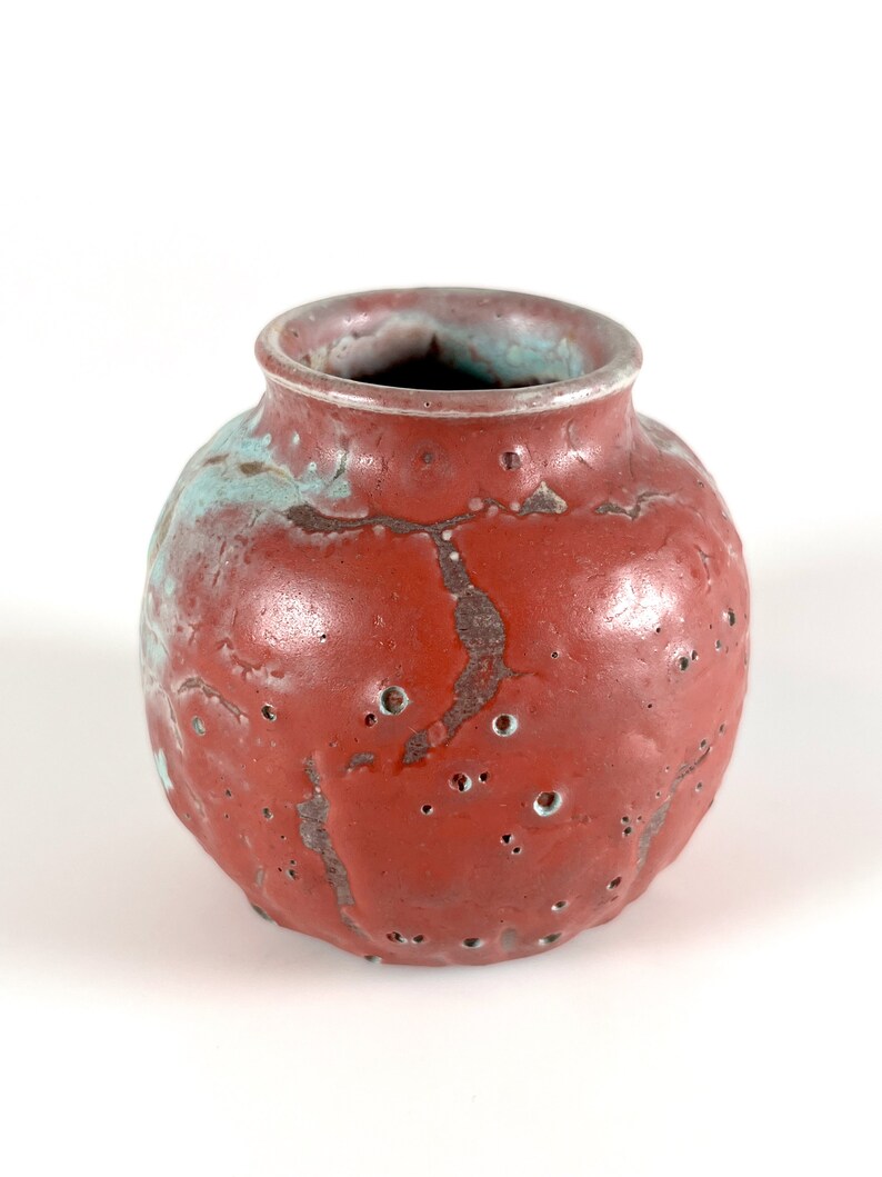 May include: A small, round, red ceramic vase with a textured surface and a light blue glaze. The vase has a wide opening and is ideal for displaying small flowers or plants.