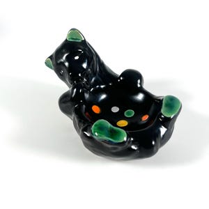May include: A glossy black ceramic bear-shaped dish with green accents on the ears and paws. The center of the dish features small, colorful circular spots in orange, yellow, and silver, creating a decorative pattern within the bowl area.