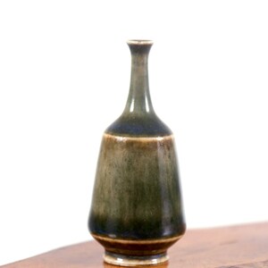 Puede incluir: Un peque&ntilde;o jarr&oacute;n de cer&aacute;mica verde oscuro con un borde marr&oacute;n y un esmalte azul en el cuello. El jarr&oacute;n est&aacute; sobre una superficie de madera.