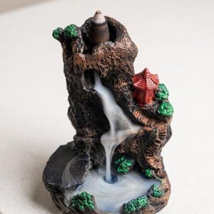 May include: A brown, rock-textured incense burner shaped like a mountain with a small red pagoda, green foliage accents, and a circular base. A lit incense cone sits at the top, releasing white smoke that flows downward like a waterfall into a pool.