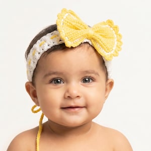 Puede incluir: Un bebé con una diadema de ganchillo hecha a mano con un gran lazo amarillo. La diadema es blanca con detalles amarillos. El bebé tiene el pelo oscuro y está sonriendo. El fondo es blanco liso.