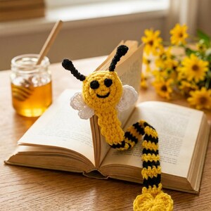 May include: A crocheted bee bookmark featuring a yellow and black striped body, a round yellow face with a smiling expression, two black antennae, and white wings. The long, flexible body extends from the top of an open book to a heart-shaped end.