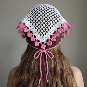 May include: A white crochet headscarf featuring a diamond-shaped openwork pattern. The edges are decorated with small, pink, three-petaled flower motifs. The headscarf is secured at the back of the head with thin, pink, crocheted tie strings.