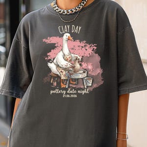 May include: A charcoal gray t-shirt featuring a graphic of a white goose wearing an apron and working at a pottery wheel. The text reads CLAY DAY and pottery date night 21.06.2026. The design includes a pink watercolor-style background splash.