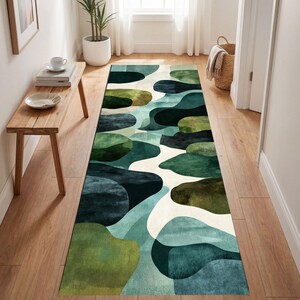 Op de afbeelding: Een lange, rechthoekige vloerkleed met een abstract ontwerp in tinten groen, blauwgroen en gebroken wit. Het patroon bestaat uit overlappende organische vormen, wat een vloeiende, moderne esthetiek creëert. Het vloerkleed ligt in een gang met houten vloer.