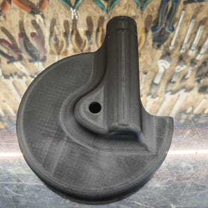 Peut inclure: Une pièce mécanique imprimée en 3D, de couleur gris foncé, avec une base circulaire et une extension cylindrique allongée. La pièce comporte un trou et un design incurvé. L'arrière-plan montre des outils.