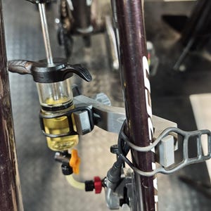 Può includere: Un kit di spurgo per freni idraulici da bicicletta fissato a un telaio di colore viola scuro. Il kit include un serbatoio in plastica trasparente riempito di liquido giallo, una maniglia a stantuffo nera, una piccola valvola arancione e un tubo.