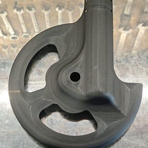 Peut inclure: Un composant noir mat imprimé en 3D présentant un bord extérieur circulaire avec deux grandes découpes, une ouverture centrale circulaire et un point de fixation cylindrique vertical au sommet avec de petits trous sur le côté du tube.