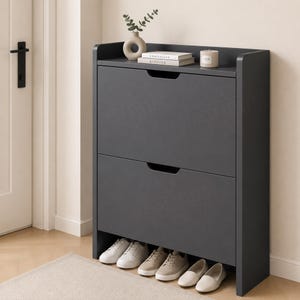May include: A dark grey shoe cabinet featuring two tilt-out storage compartments with cutout handles. The top surface holds a beige ceramic vase with a small plant, two stacked books titled Kinfolk and Christiane, and a small cylindrical candle.