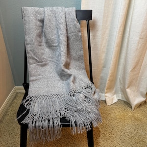 May include: A light gray rectangular scarf with a soft, fuzzy texture. The scarf features a decorative woven border at the bottom edge, which transitions into long, loose, matching gray fringe tassels that hang down from the hem.