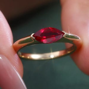 May include: A gold ring featuring a single marquise-cut deep red gemstone set horizontally in a smooth metal band. The setting holds the stone securely with two prongs at the pointed ends, creating a clean and polished look for the jewellery piece.