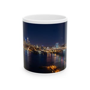 Peut inclure: Mug en céramique blanche avec une vue nocturne d'une ville, un pont et des lumières se reflétant sur l'eau. Le mug a un fond bleu foncé et un intérieur blanc.