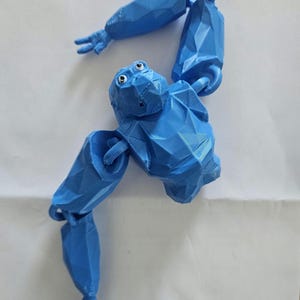 May include: A bright blue, low-poly articulated gorilla figure with a geometric, faceted surface texture. The figure features a head with white and black eyes, a torso, and two long, jointed arms with three-fingered hands, all constructed in a single color.