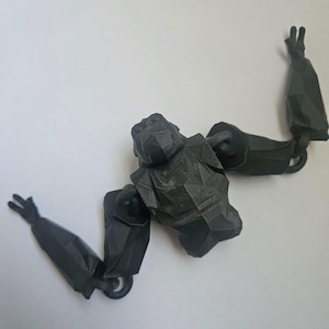 May include: A matte black, low-poly style gorilla figurine featuring a geometric, faceted surface texture. The figure has a central torso and head section with two long, articulated arms extended outward, each ending in a three-fingered hand.