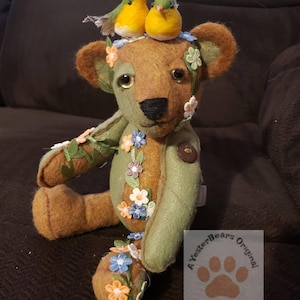 May include: A handcrafted teddy bear with a brown and green color scheme, adorned with floral accents and two small bird figurines perched on its head. The bear has a button on its chest and a tag that reads "A YesterBears Original".
