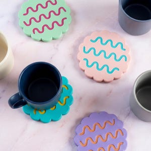 Puede incluir: Un juego de cuatro posavasos redondos con bordes festoneados en verde menta, rosa claro, azul cerceta y morado, cada uno con un patrón de líneas onduladas en contraste. Una taza azul oscuro descansa sobre el posavasos cerceta junto a otras tres tazas.