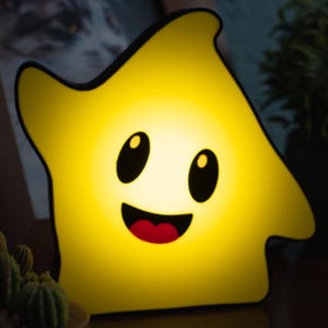 Puede incluir: Una luz de noche amarilla en forma de estrella con una cara sonriente. La luz tiene ojos negros y una boca roja. Los bordes de la luz están delineados en negro. La luz brilla intensamente.
