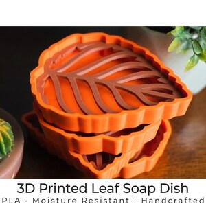 Puede incluir: Una pila de tres jaboneras con forma de hoja, impresas en 3D, de color naranja. El diseño de la hoja presenta vetas marrones y el plato tiene un borde texturizado. El texto en la parte inferior dice "3D Printed Leaf Soap Dish".