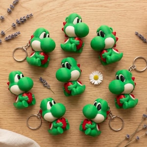 Puede incluir: Llaveros de Yoshi verdes y blancos, cada uno sosteniendo un corazón rojo, dispuestos sobre una superficie de madera clara. Los llaveros tienen una apariencia impresa en 3D. También hay una pequeña margarita y ramitas de lavanda.