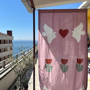 May include: A pink curtain adorned with white dove and red heart motifs. The curtain also features red tulip designs with green stems and leaves. It hangs outdoors on a sunny day, with a view of the sea and buildings in the background.