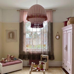 Op de afbeelding: Een heteluchtballonvormige hanglamp met roze en witte strepen en donkerrode kwastjes. Het raam heeft een bruin, doorschijnend gordijn met een witte zwaan, twee gele tulpen en de naam Sophia in het wit geborduurd op de stof.