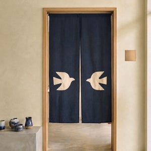 Può includere: Una tenda noren in tessuto blu navy scuro appesa a una porta in legno. La tenda è divisa in due pannelli, ognuno dei quali presenta una grande sagoma di uccello color crema rivolta verso il centro. Il tessuto ha una consistenza visibile.