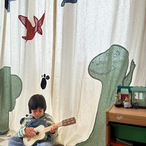Pode incluir: Uma criança toca ukulele branco num chão xadrez. O fundo apresenta uma cortina com ilustrações de dinossauros e pássaros em verde, vermelho e azul. Um armário de madeira com brinquedos está à direita.