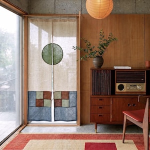 Op de afbeelding: Een beige linnen deurgordijn met een gespleten groen cirkelontwerp aan de bovenkant en een patchworkpatroon van blauwe, bruine en groene vierkanten aan de onderkant. Het gordijn hangt in een houten frame naast een houten kast met een vintage radio.