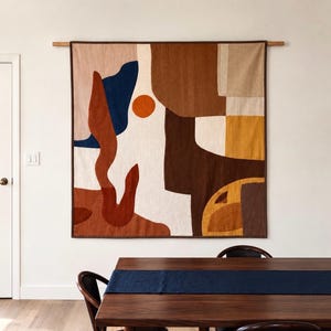 May include: A large rectangular fabric wall hanging featuring an abstract composition of geometric and organic shapes in shades of cream, terracotta, navy blue, dark brown, and mustard yellow, suspended from a horizontal wooden rod.