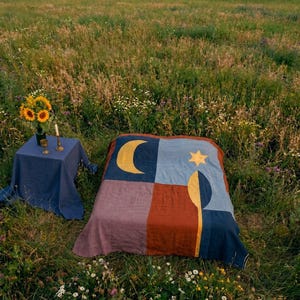 May include: A rectangular blanket featuring a color-blocked design in shades of blue, rust, and mauve, adorned with a yellow crescent moon, a yellow star, and a yellow vertical leaf motif. Beside it, a blue-covered table holds a vase of sunflowers.