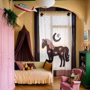 Puede incluir: Una habitación con una cortina grande que presenta un gráfico de caballo marrón oscuro, estrellas rosas y una luna creciente. El espacio incluye un diván amarillo claro con falda con volantes, un dosel marrón oscuro, un sillón y un armario rosas.