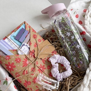 Op de afbeelding: Een geschenkdoos met een doorzichtige waterfles met lavendelbloemenprint en een lichtroze deksel, een bruin papieren pakketje met rode bloemenpatronen vastgebonden met touw, een lichtpaarse scrunchie met stippen en een doorzichtige plastic haarklem.