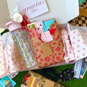 Peut inclure: Une boîte en carton blanc contenant une gourde transparente à motifs floraux, un cadeau emballé dans du papier kraft à fleurs rouges, un stylo rose, un sachet de thé, une pince à cheveux noire, un chouchou noir et une carte avec le texte gracias BooksBox.