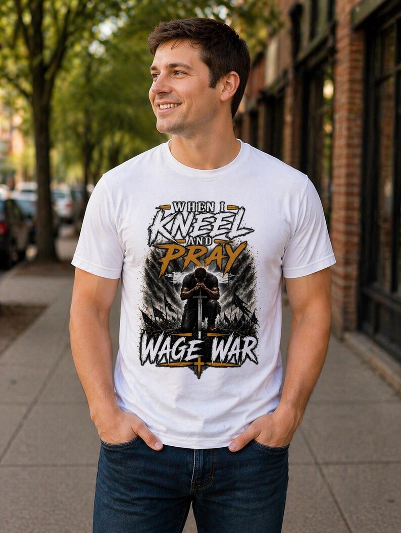 May include: White t-shirt with the text "WHEN I KNEEL AND PRAY WAGE WAR" in a stylized design. The graphic features a kneeling figure with a sword. The shirt is being worn by a person in blue jeans.