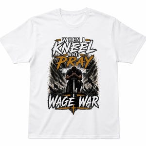 May include: White t-shirt featuring the text "WHEN I KNEEL AND PRAY I WAGE WAR" in a stylized design. The graphic includes a kneeling figure with a sword, set against a backdrop of flags and a cross.